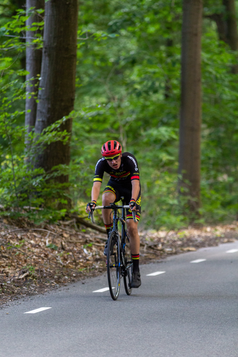 A man wearing a black and red jersey on a bicycle riding down a road with trees in the background.