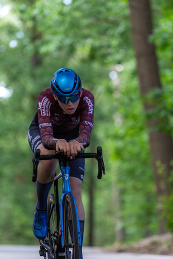 A bicycle racer wearing blue and black bike gear, is riding a bicycle down the road in front of trees.