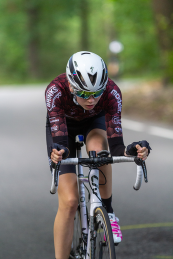 A woman in a red jersey is riding her bike down a street. She has on a white helmet and sunglasses for protection.