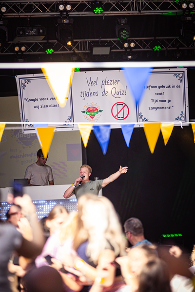 A man is holding a microphone while pointing with his right hand. He is wearing a shirt that says "Pubquiz".