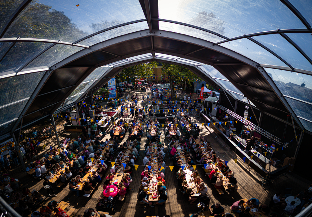 A crowd of people gathers around tables in a large tent with an open roof.