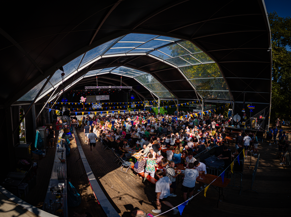 A group of people sit in chairs and tables under a tent with signs that say Pubquiz.