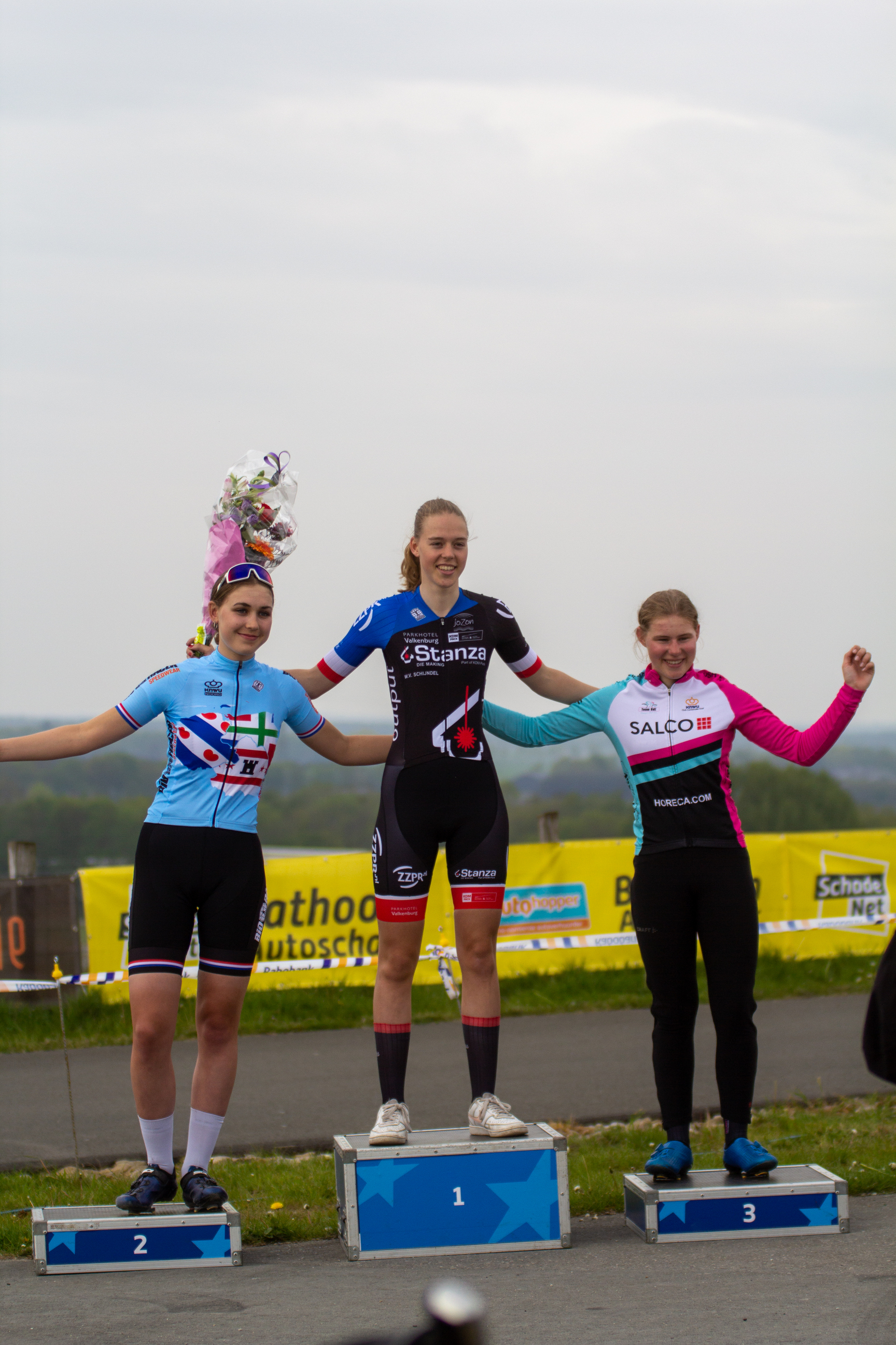 Three female cyclists stand on podiums at the Coll du Vam Tweedaagse race.