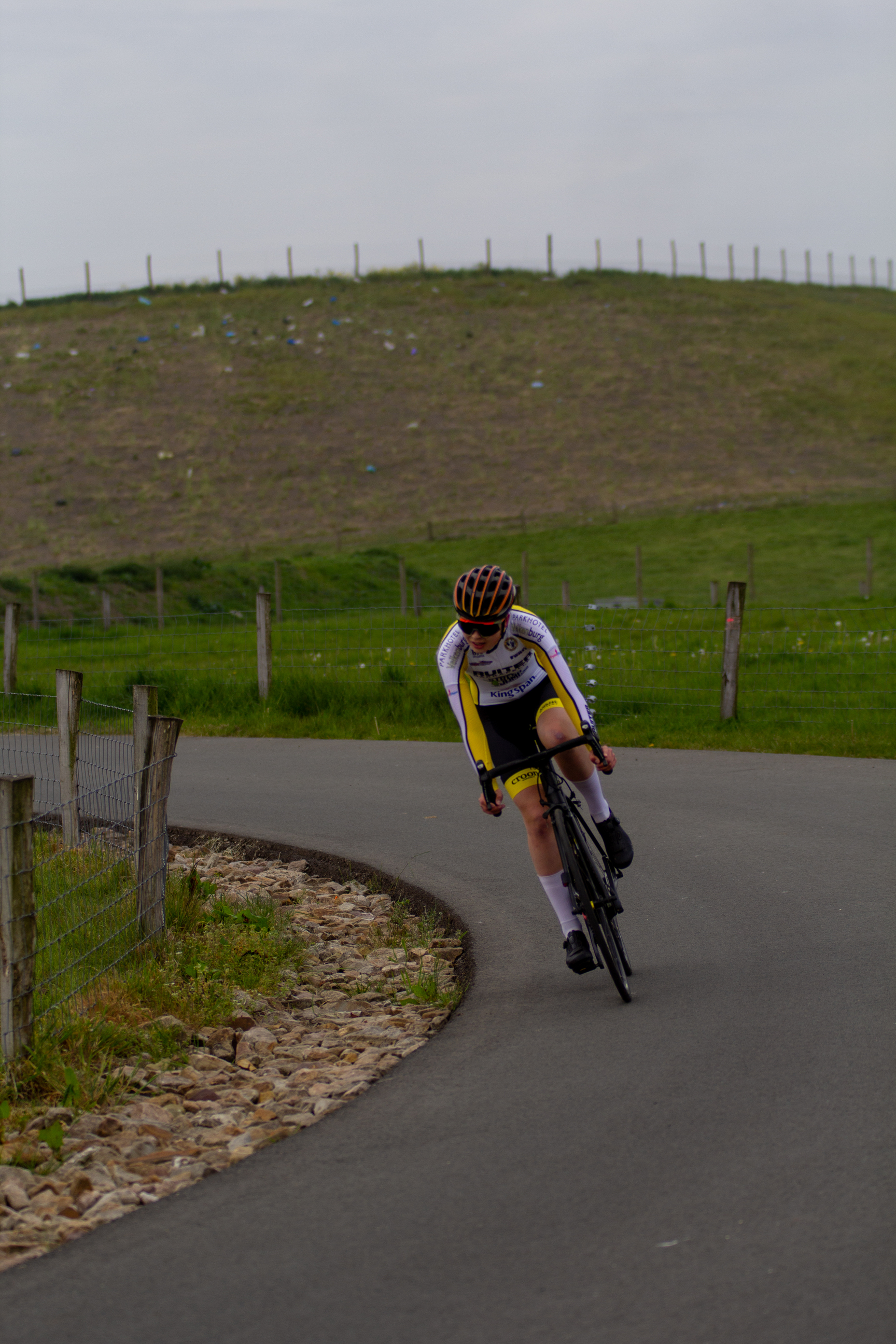 A woman in a yellow and white cycling outfit rides her bicycle down a hillside road with grass on either side.