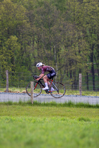 A female cyclist with the number 13 on her jersey rides down a rural road on a sunny day.