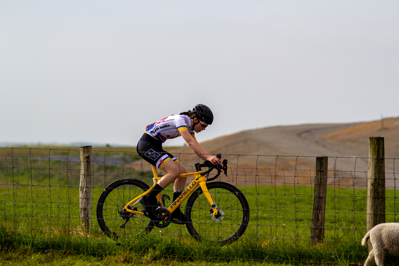 A cyclist wearing a black outfit is on the road, riding a yellow bike. She is looking at the ground as she pedals along.