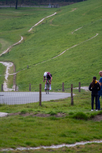 Three people stand on the side of a road, watching as a man rides his bike nearby.