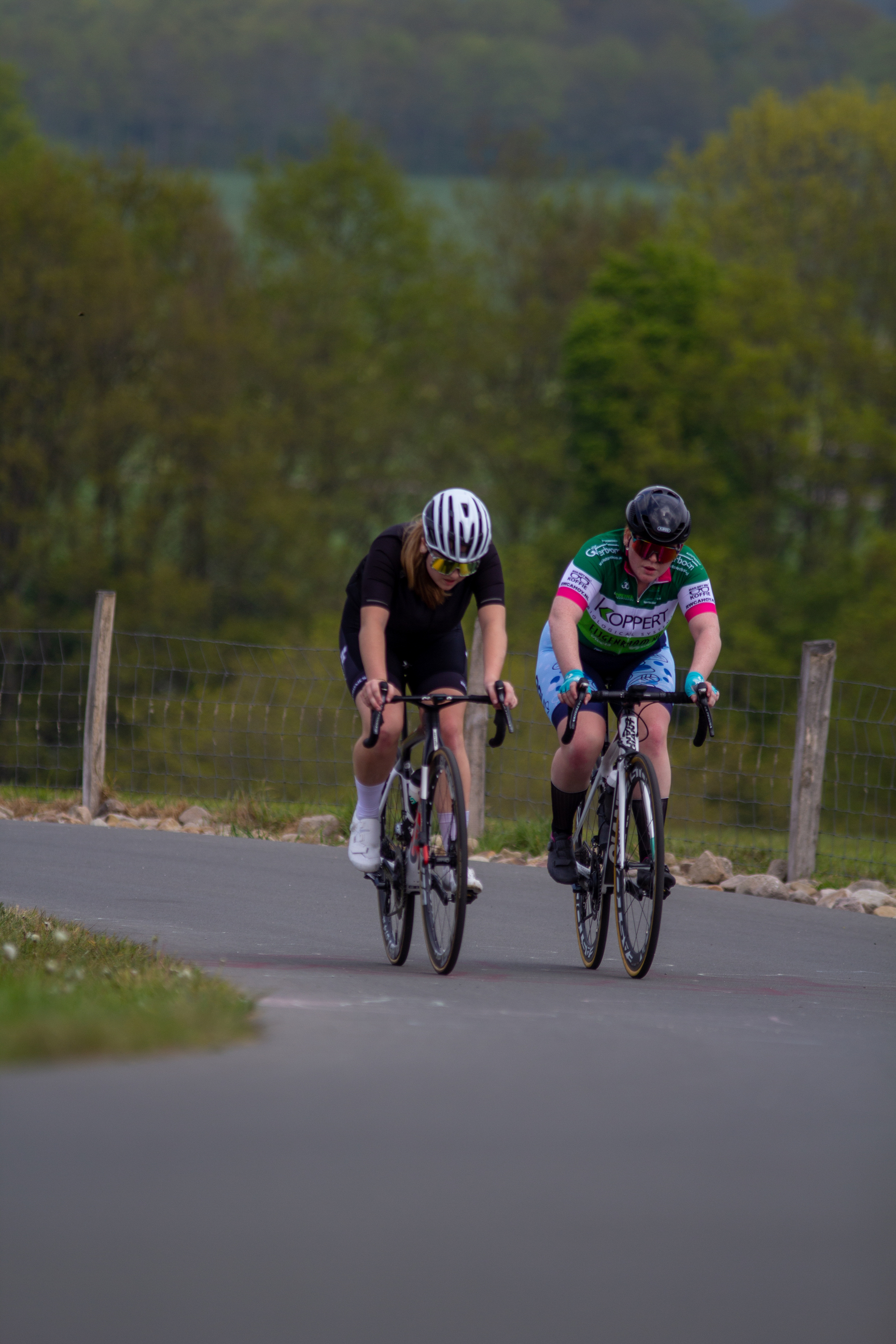 Two women are riding bikes on a road. They both have helmets and one has a pink shirt with the word "Vlaenderen" across it.