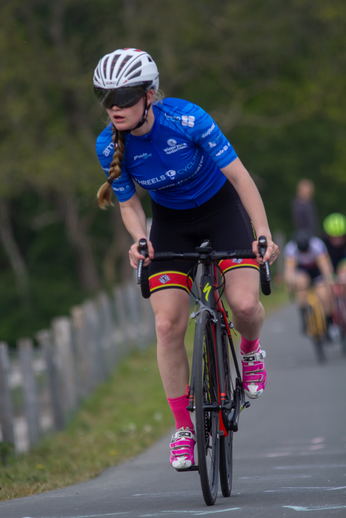 A woman in a blue shirt and a black helmet rides her bike on a road. She is number 1 in the women's race of a cycling event.