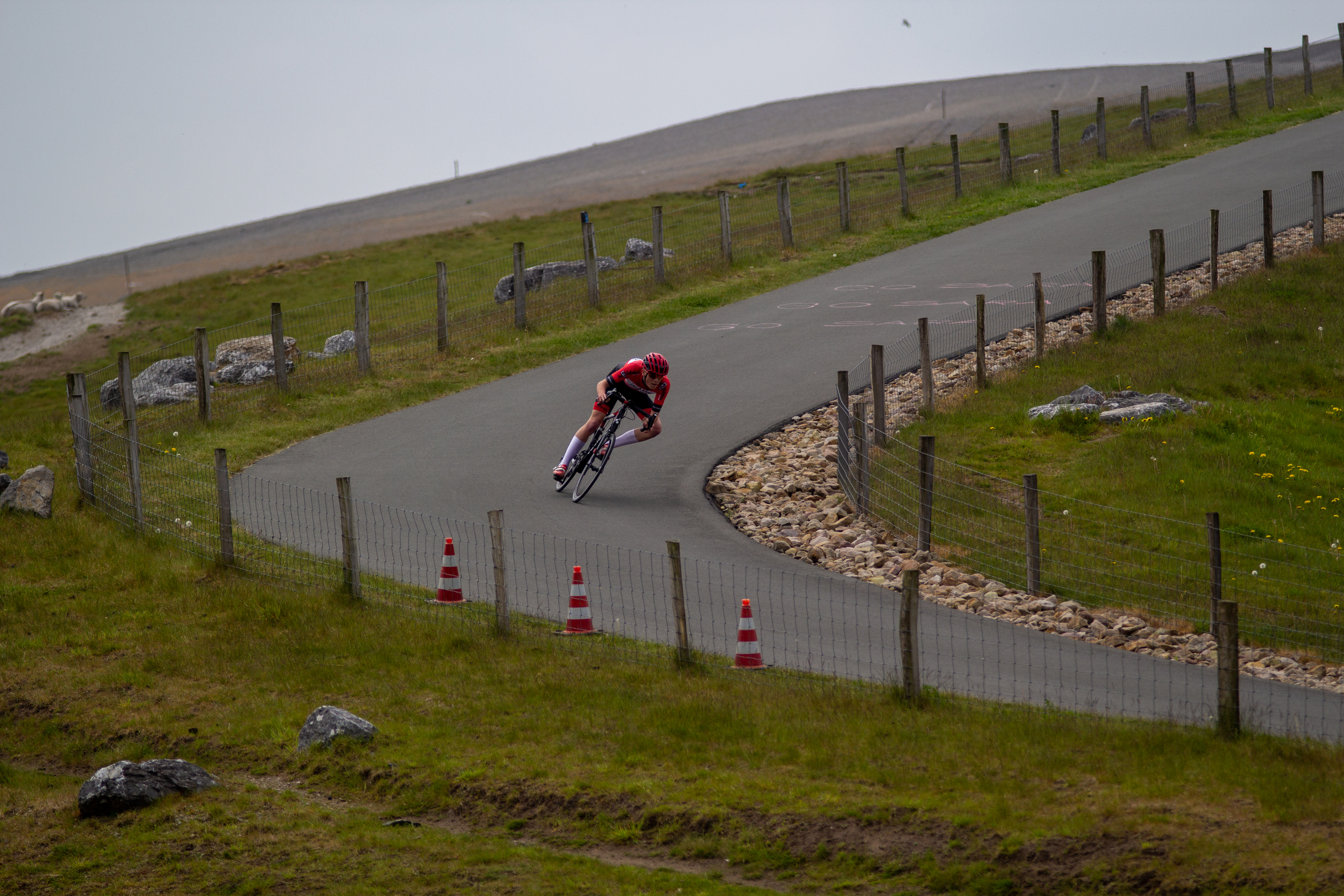 A cyclist in a red and black outfit races on a track with orange and white flags.