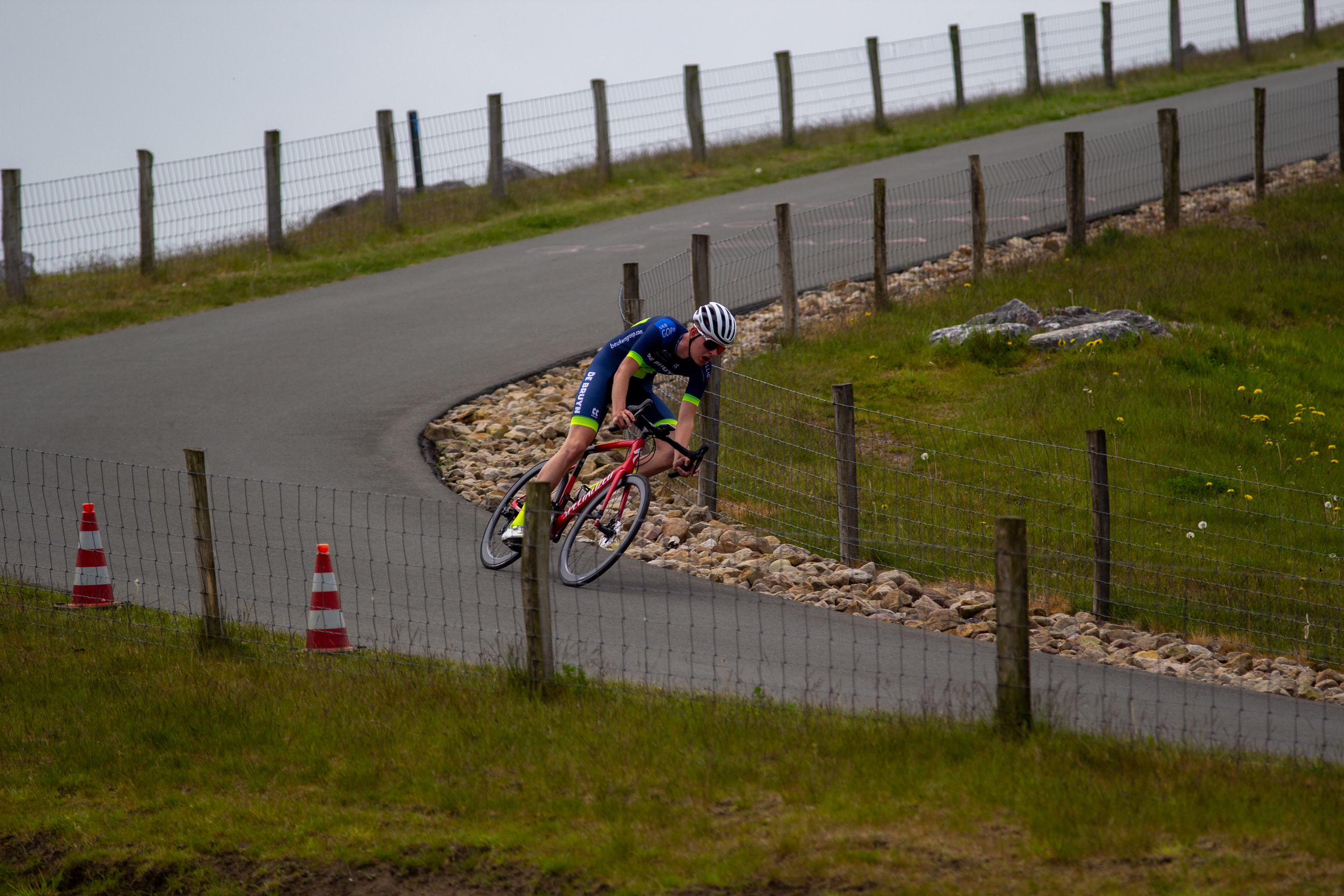 A bicyclist races down a road, wearing blue and green and heading towards a red cone.