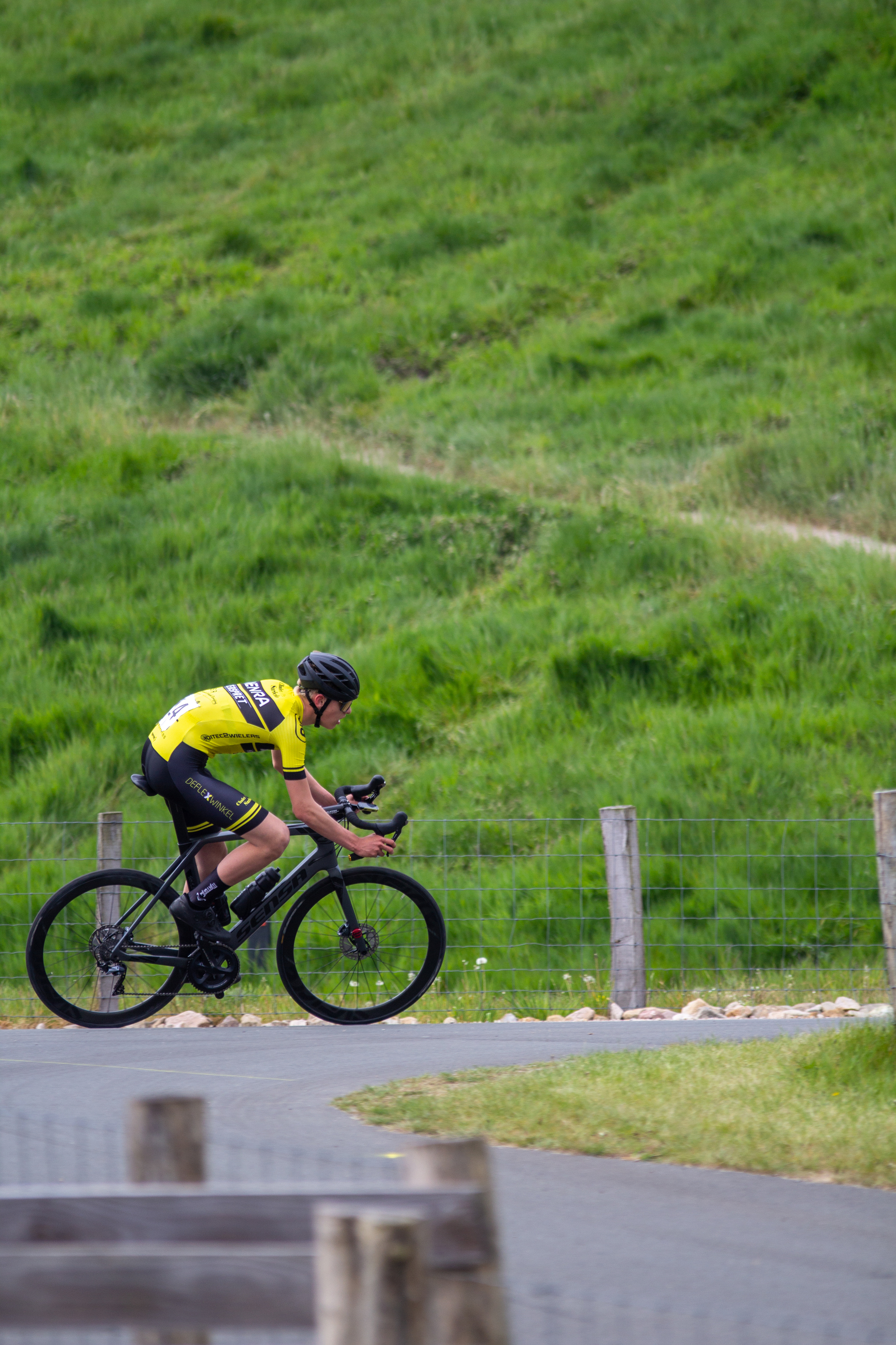 A cyclist riding a bike on a road with grass and hills in the background.