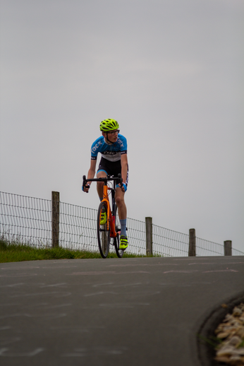 A woman in a blue and white jersey, along with a yellow helmet is riding her bicycle down the road.