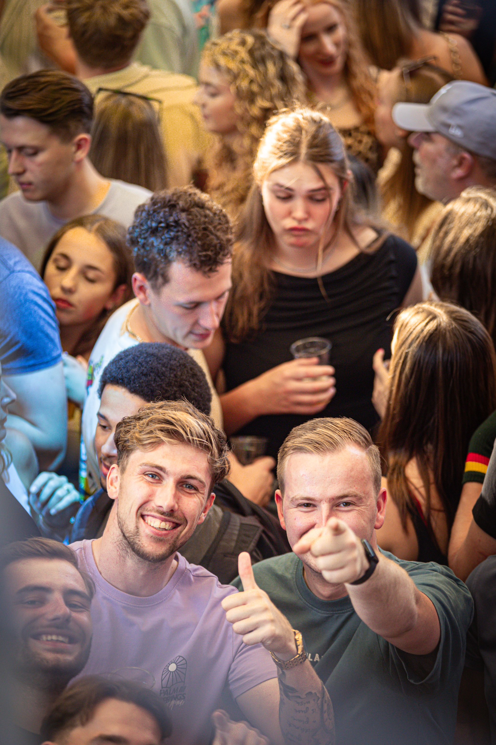 A man sitting in a crowded place, giving a thumbs up with his right hand.