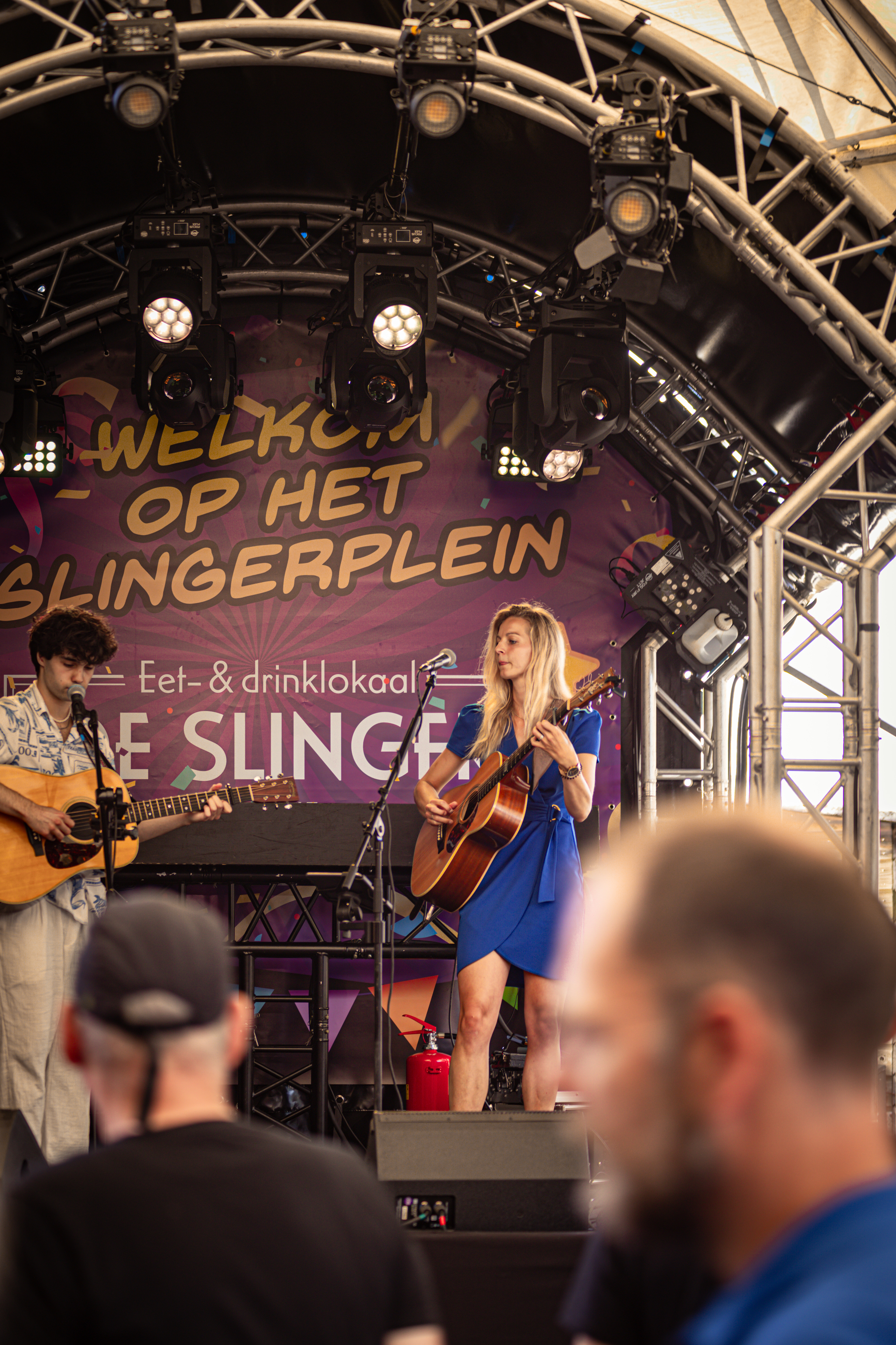 A rock band on stage in front of a sign that reads Welkom op het Slingerplien and Lee Slanger.