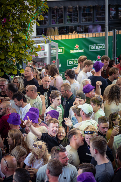 A lively outdoor gathering of people with a green backdrop that says "De Slinger" in white letters.