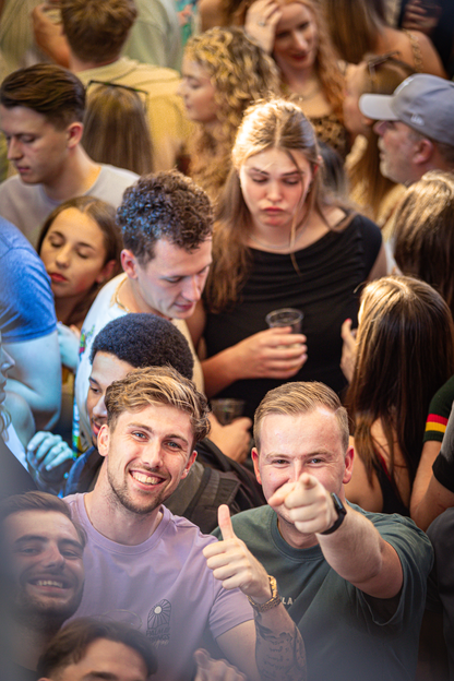 A man sitting in a crowded place, giving a thumbs up with his right hand.