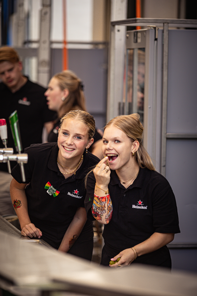 Two girls, one with a tattoo on her arm, wearing black shirts that say De Kiek.