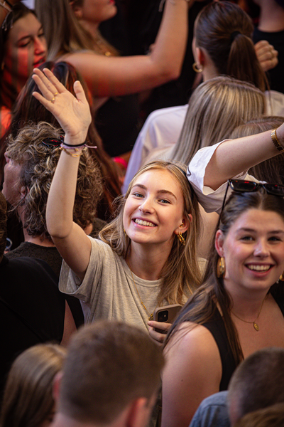 A woman in a grey shirt is smiling as she waves to the camera while standing near others.