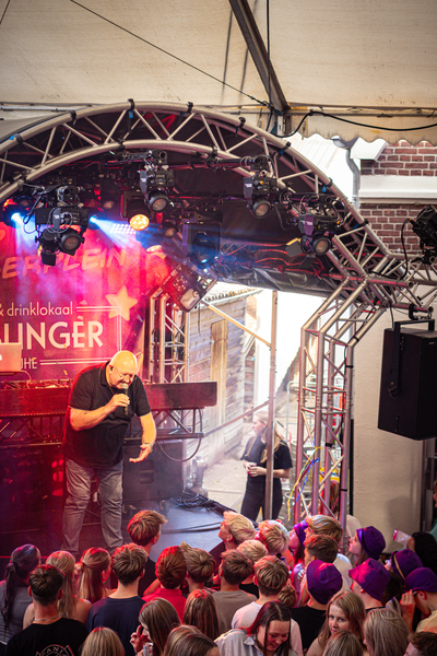 A man wearing a black shirt standing on stage next to a banner with the word "slinger" written in white.