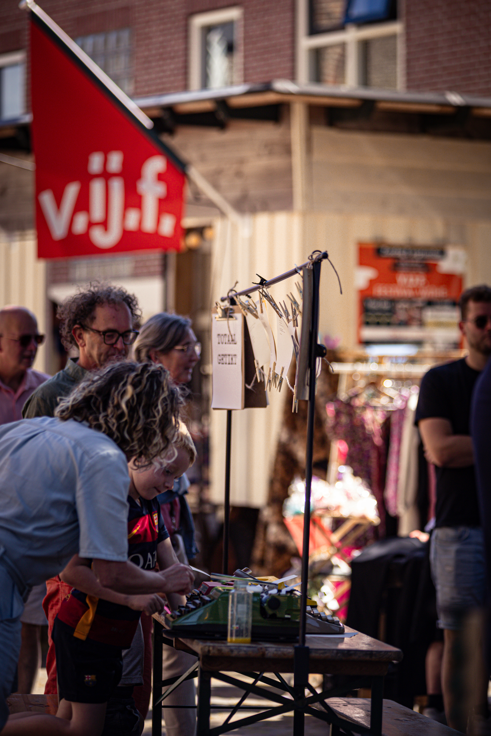 "V.IJ.F. Festival shoppers and vendors are seen around a table with clothing displayed for sale."