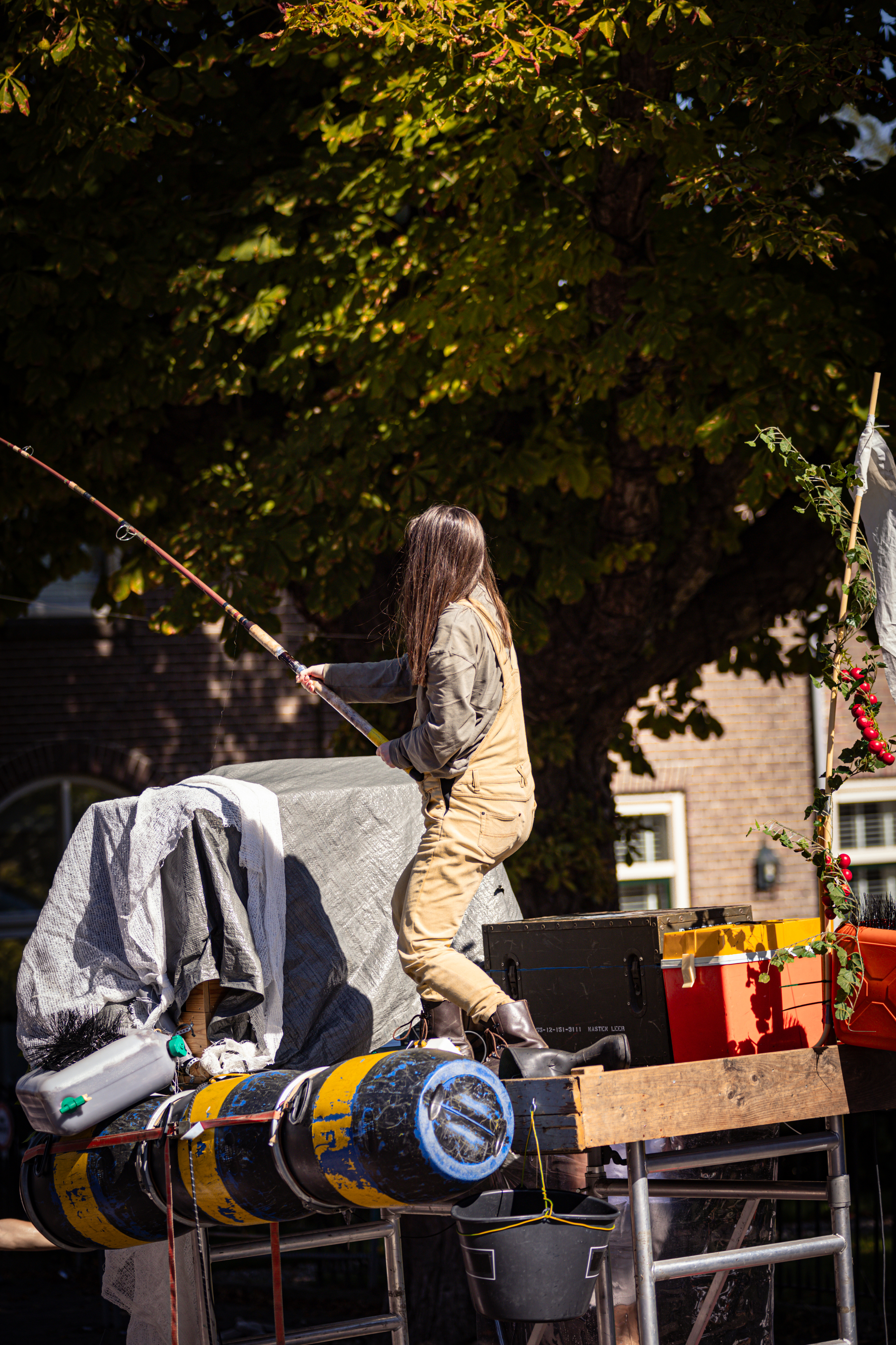 The woman in the orange jumpsuit is a performer at the V.IJ.F. Festival, where she is juggling on stilts with a fishing rod.