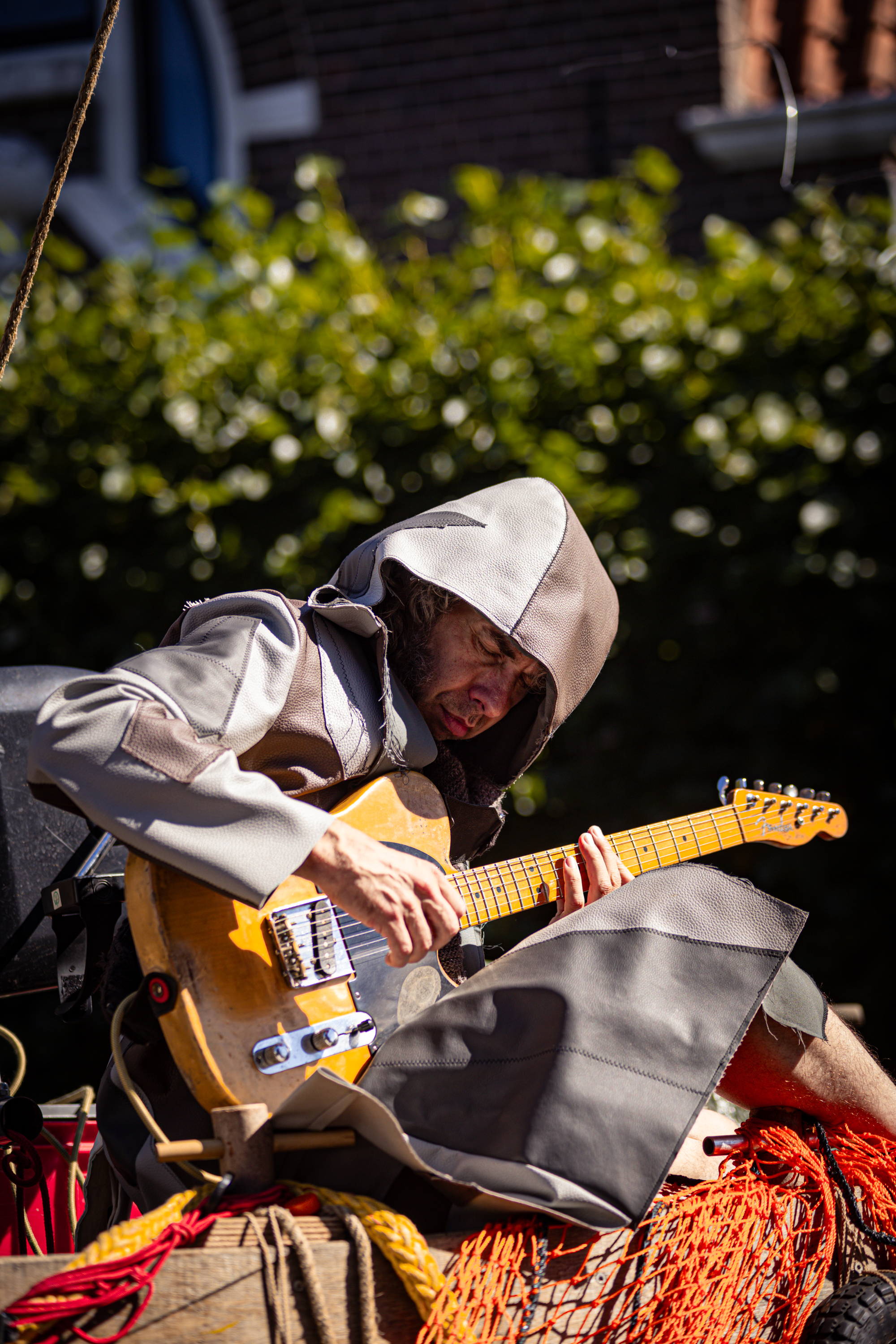 A man playing a guitar outside, wearing a hoodie and holding the neck of his instrument with his left hand.