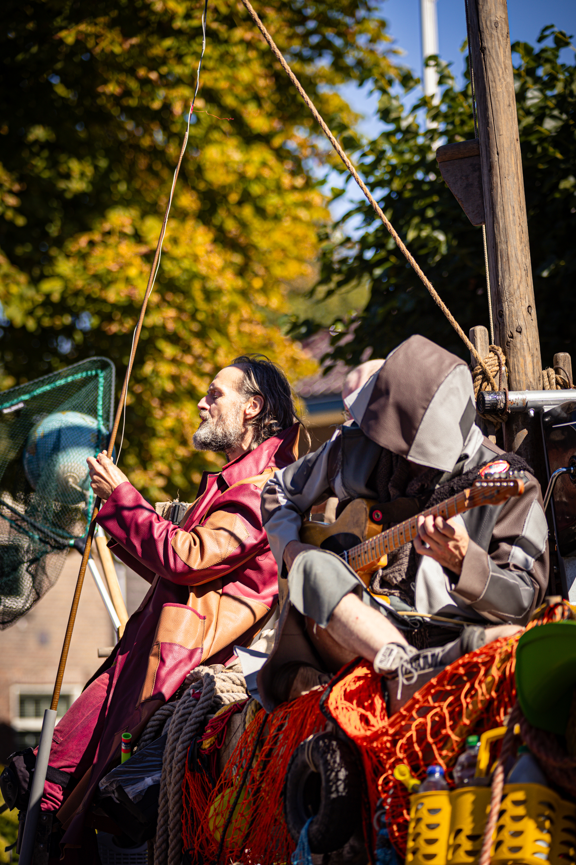 A man wearing a hood sits on a boat playing guitar in the V. IJ. F. Festival.