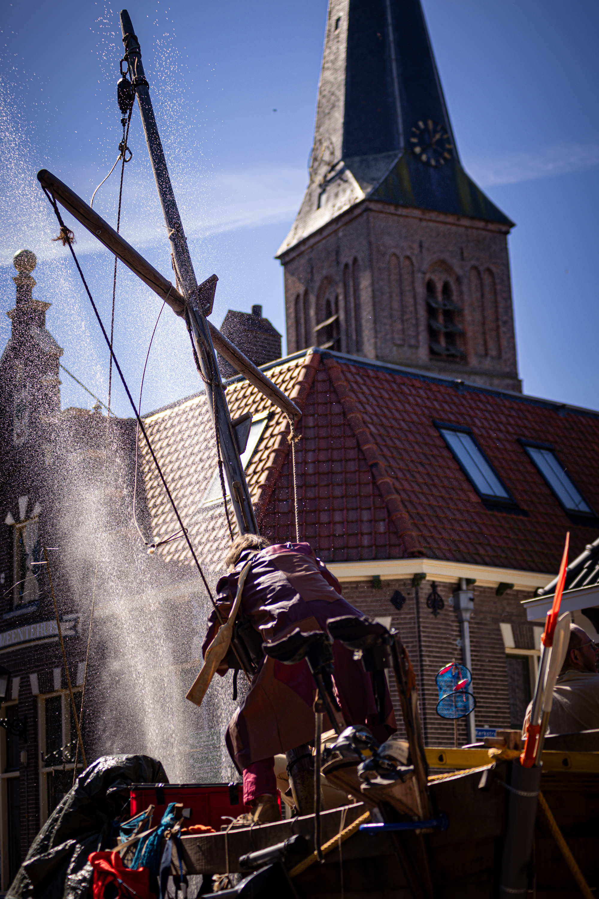 A woman standing near a water pump in the V.IJ.F. Festival, wearing a purple jacket and holding onto a rope.