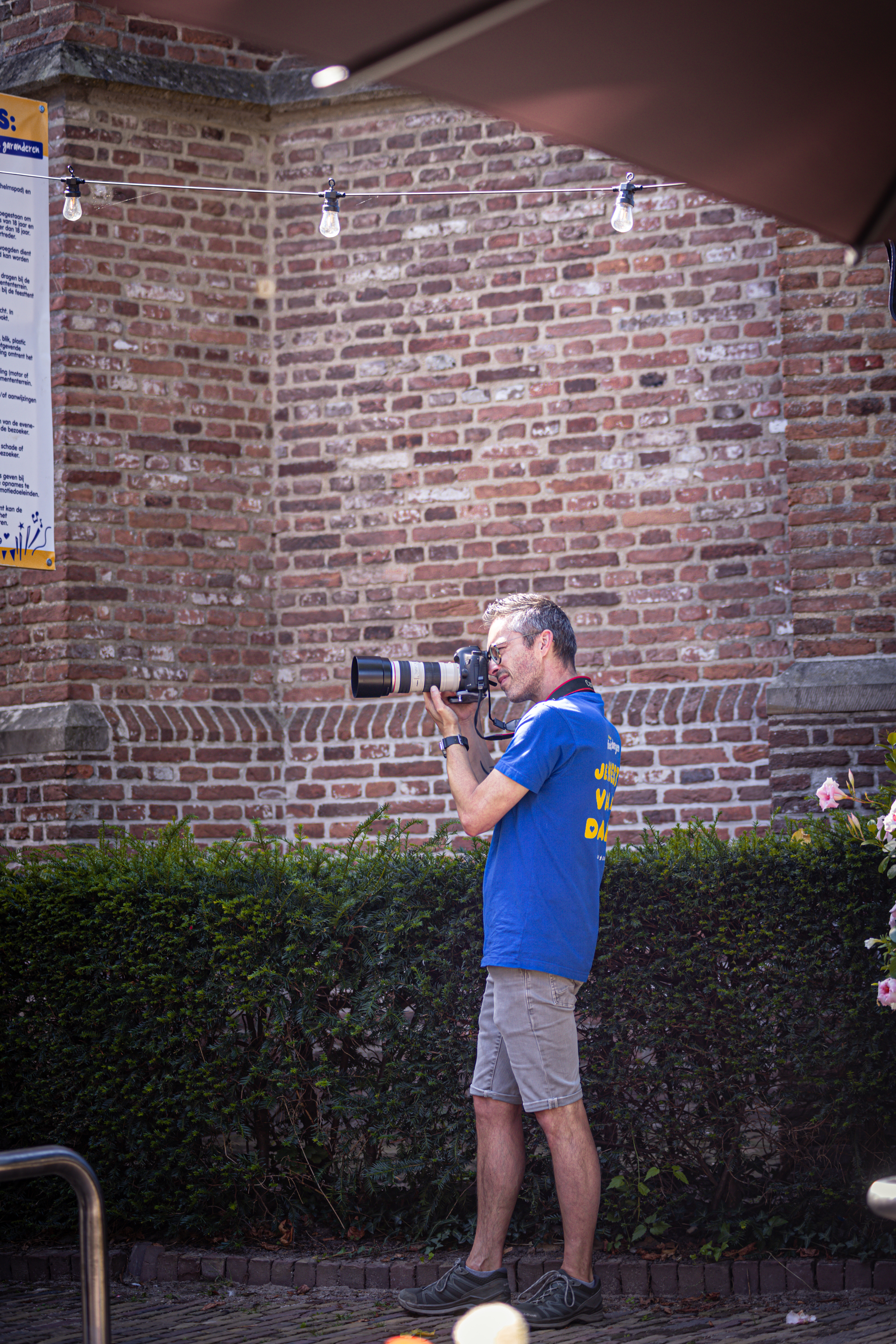 A man in a blue shirt stands outside a brick building with a camera.