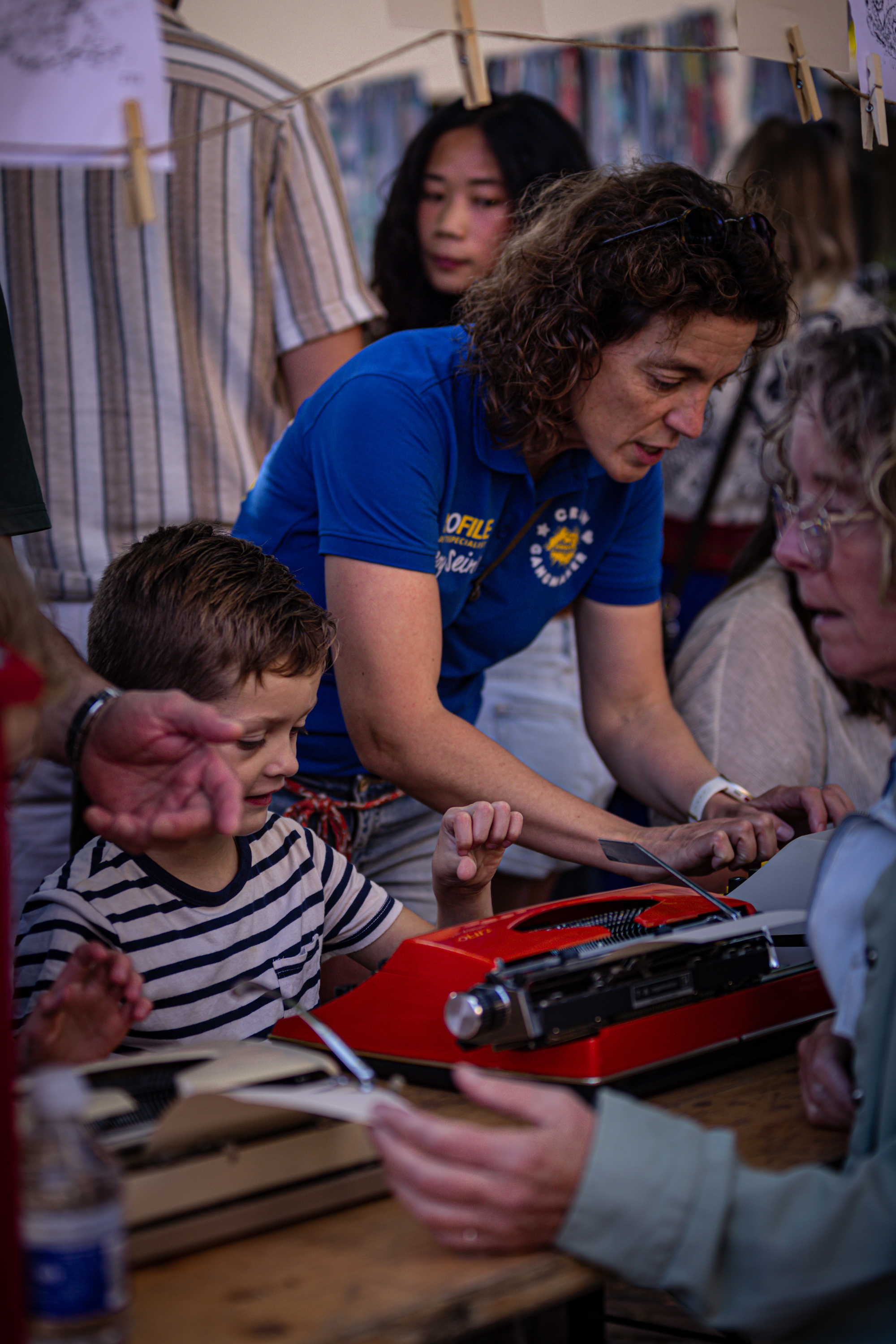 A woman in a blue shirt and a child are at a desk. The woman is showing the child something on a red typewriter.
