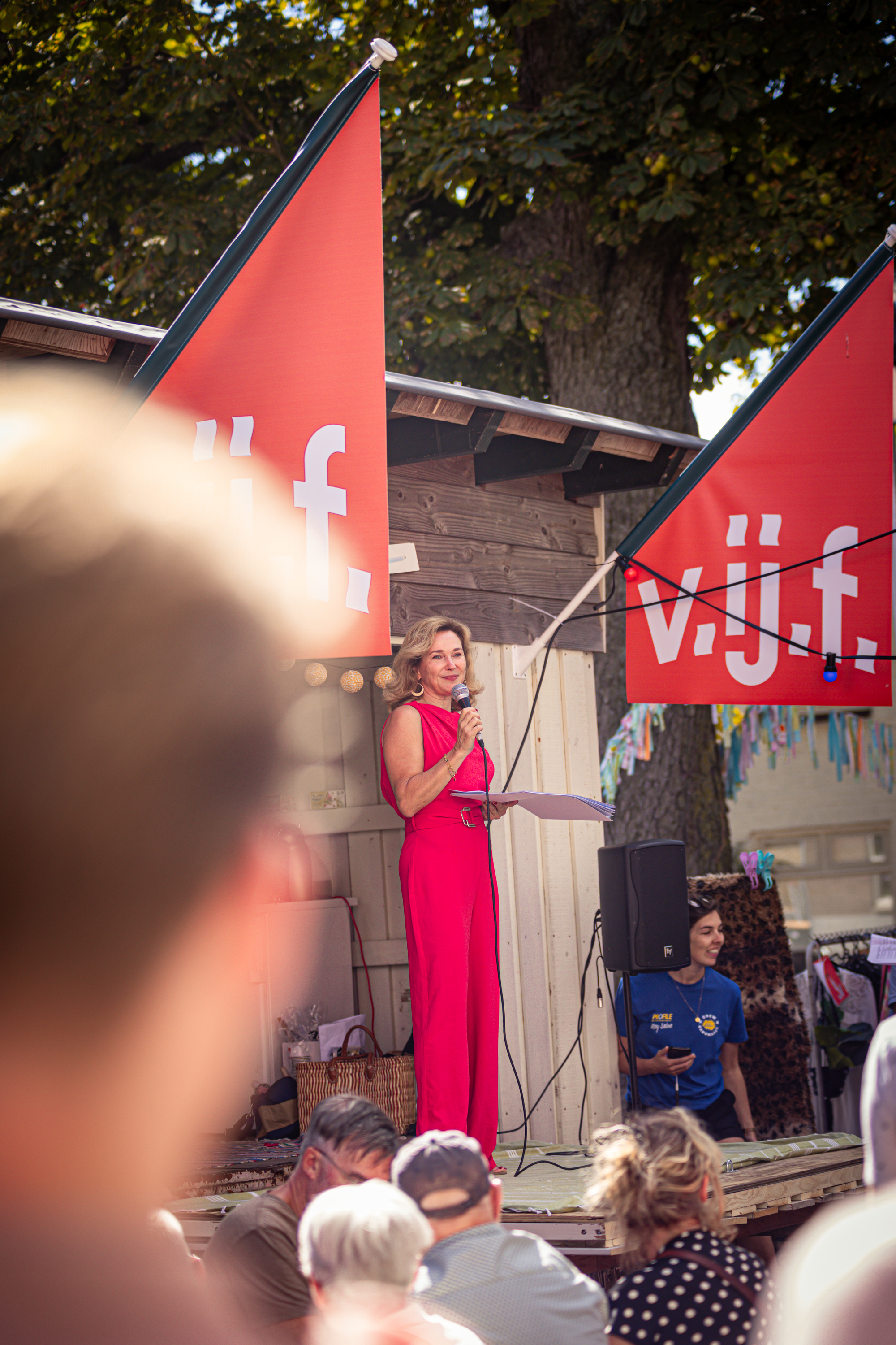 A woman is standing in front of two flags on a stage at the V.I.J.F. Festival, talking into a microphone.