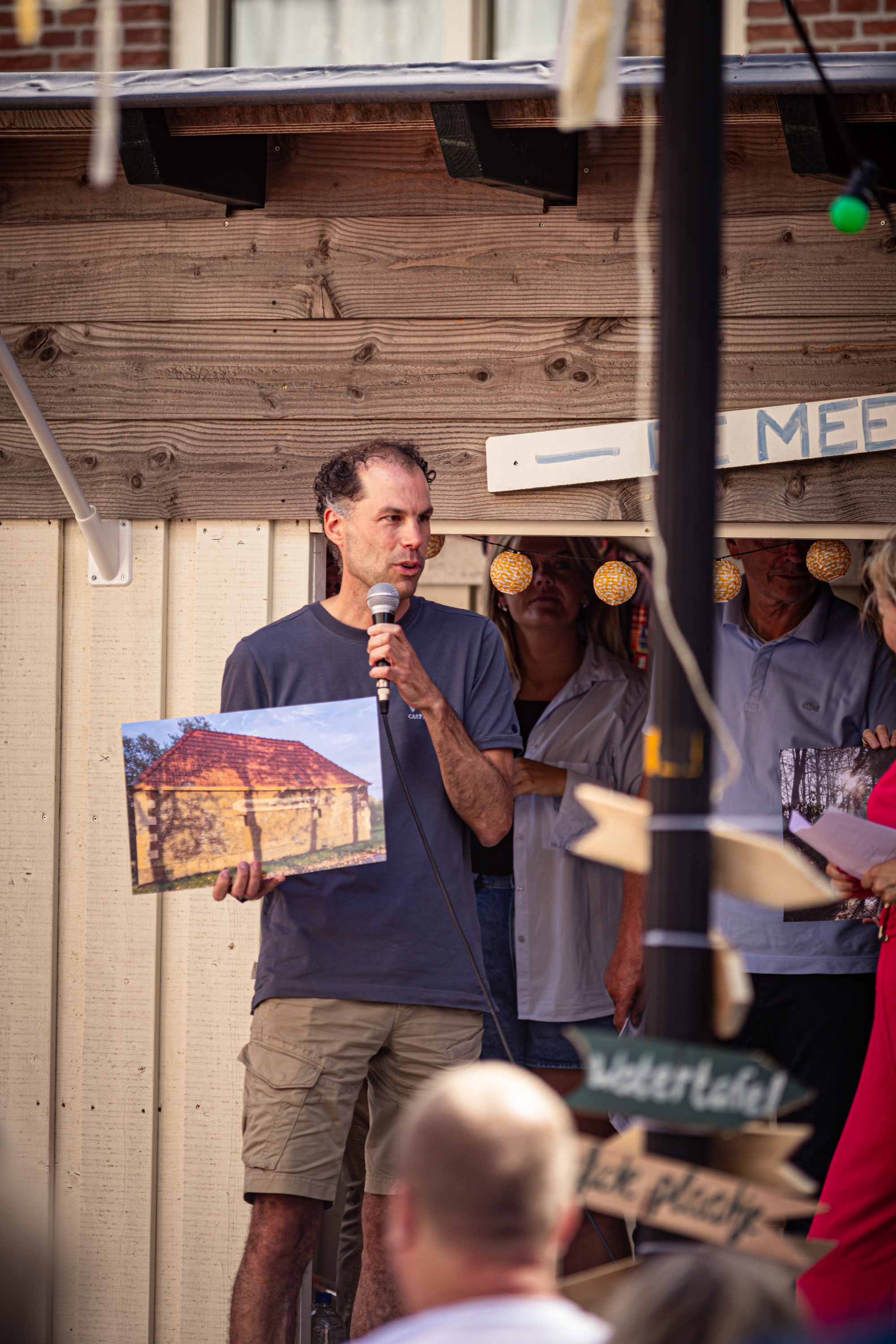 A man is speaking into a microphone at the V. I. J. F. Festival, with a sign for the Kosterij nearby.
