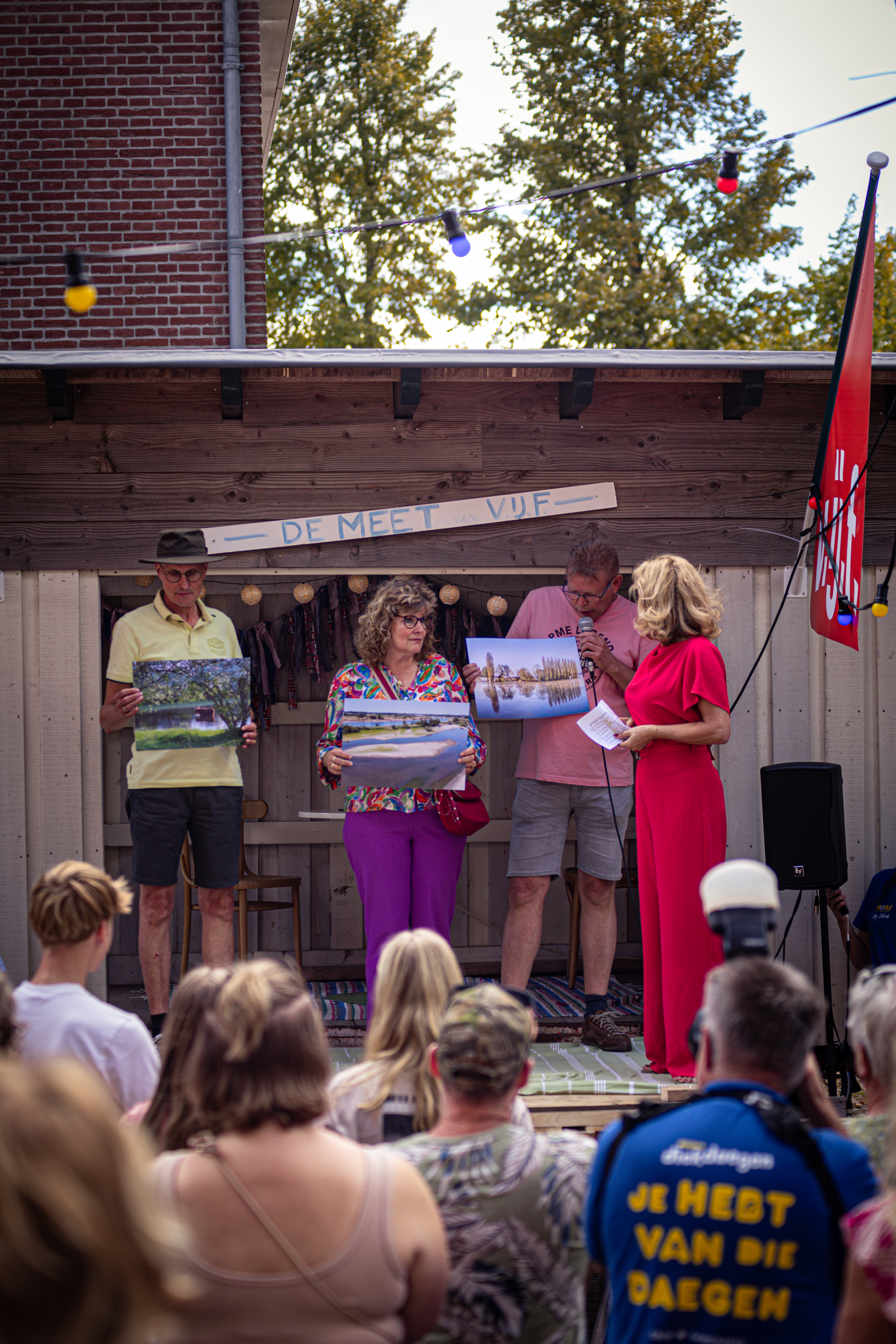 A woman is on stage at the V.IJ.F Festival, holding a photo and surrounded by people.