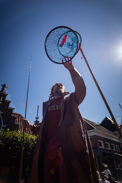 A man wearing a red coat and purple pants holding a blue circular object with red tassels above his head.