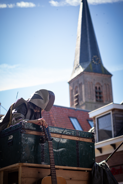 A man plays a guitar in front of a clock tower, with a green trunk behind him.
