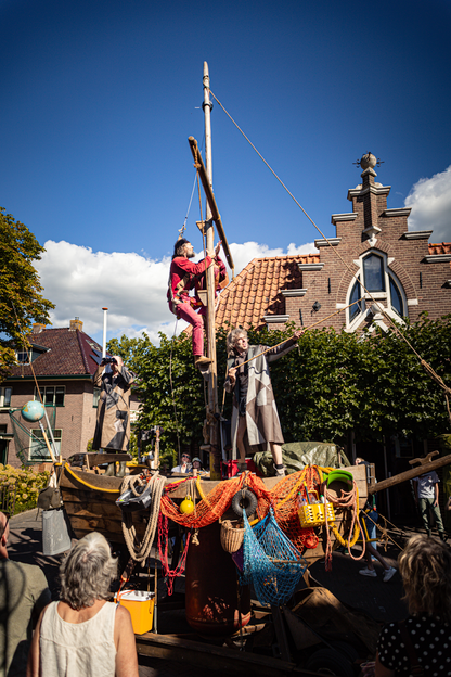 An art installation at the V.IJ.F. Festival features a man hanging from a crane, and various objects hang below him.