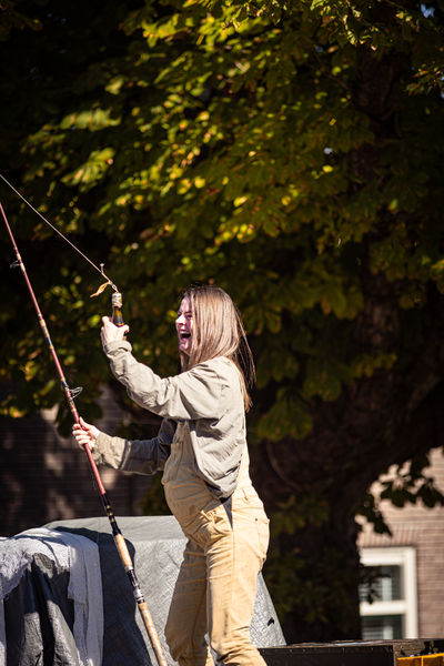 A woman is smiling while fishing. She's wearing a beige shirt and khaki pants, holding a rod in her left hand.