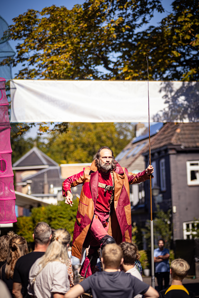 A man wearing a red cape and holding a flag stands in front of people at the V.IJ.F. Festival.