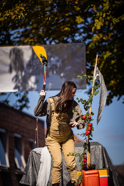 A woman wearing an apron, holding a yellow shovel. She is standing next to a potted plant at the V.I.J.F. Festival.