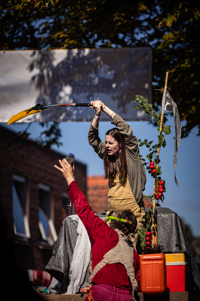 Two people in a park are making puppets with clothes and other items, including red and orange tubs.