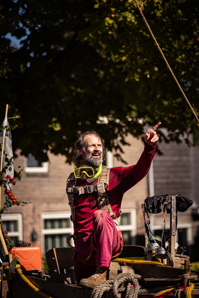 A man in a red shirt sitting on a boat. He has his hand up and is wearing goggles.