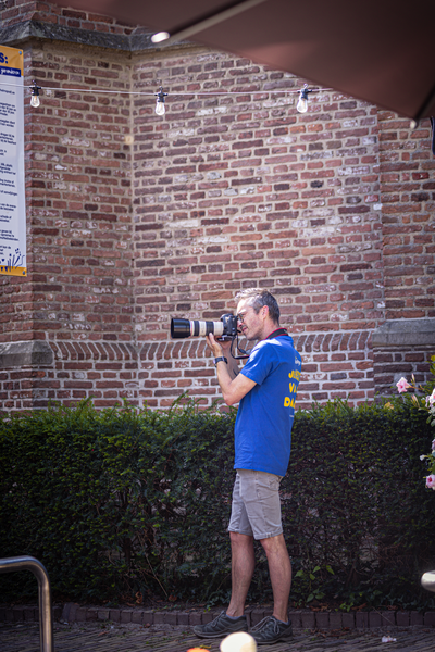 A man in a blue shirt stands outside a brick building with a camera.