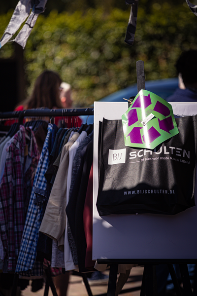 A green paper bag with a purple flower on it hangs from a metal stand. The bag is in front of other hanging clothes and bags.