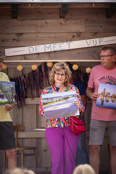 A woman is holding a painting of a village with a man next to her. They are in front of a sign that says "De Meet Vuf".