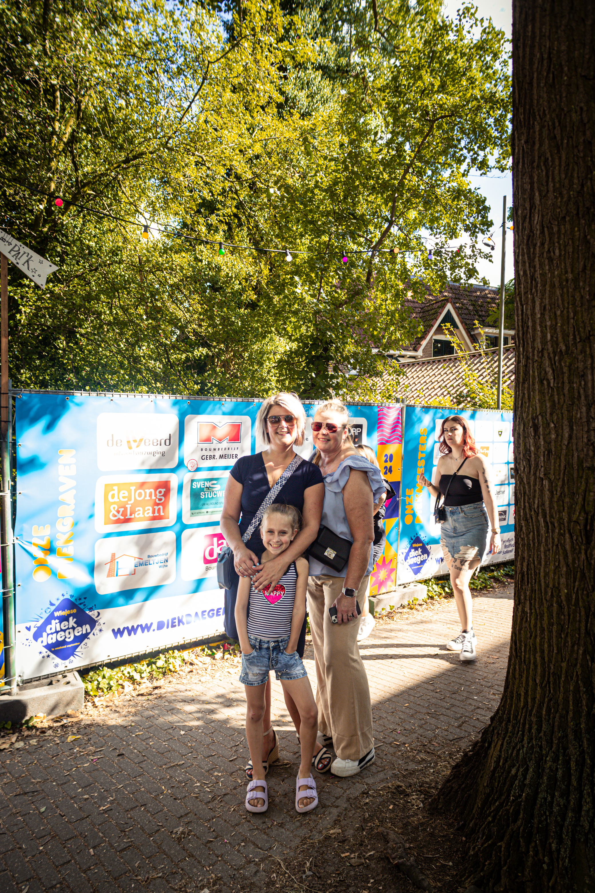 A family is posing for a photo at the entrance of a park. The park has an advertisement for Smaek in 't park on its fence.