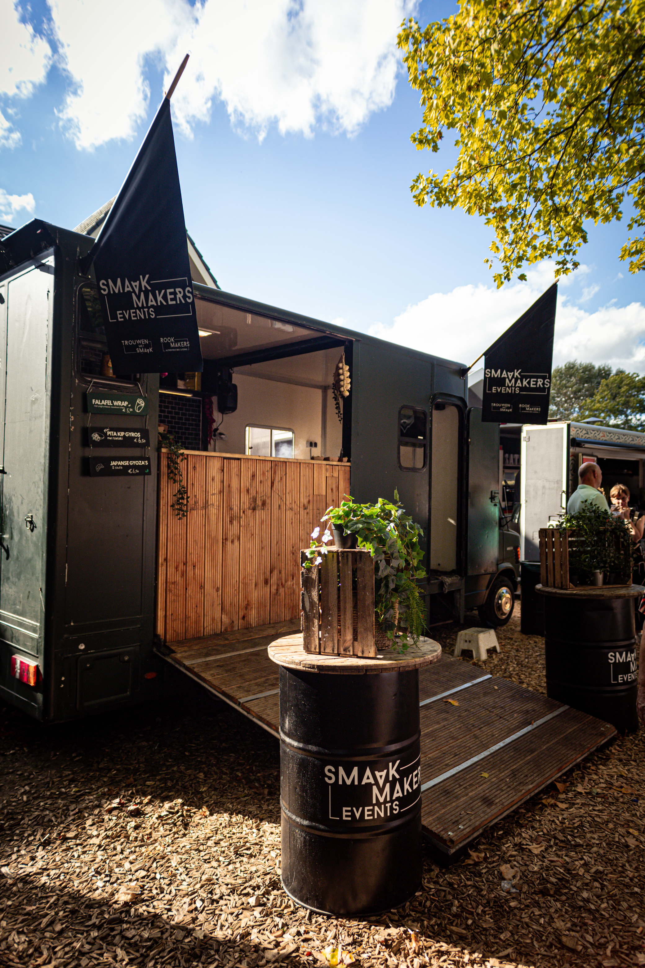 Food truck called "Smaek in 't park" serving customers. The food truck is black and has two flags hanging from the roof.