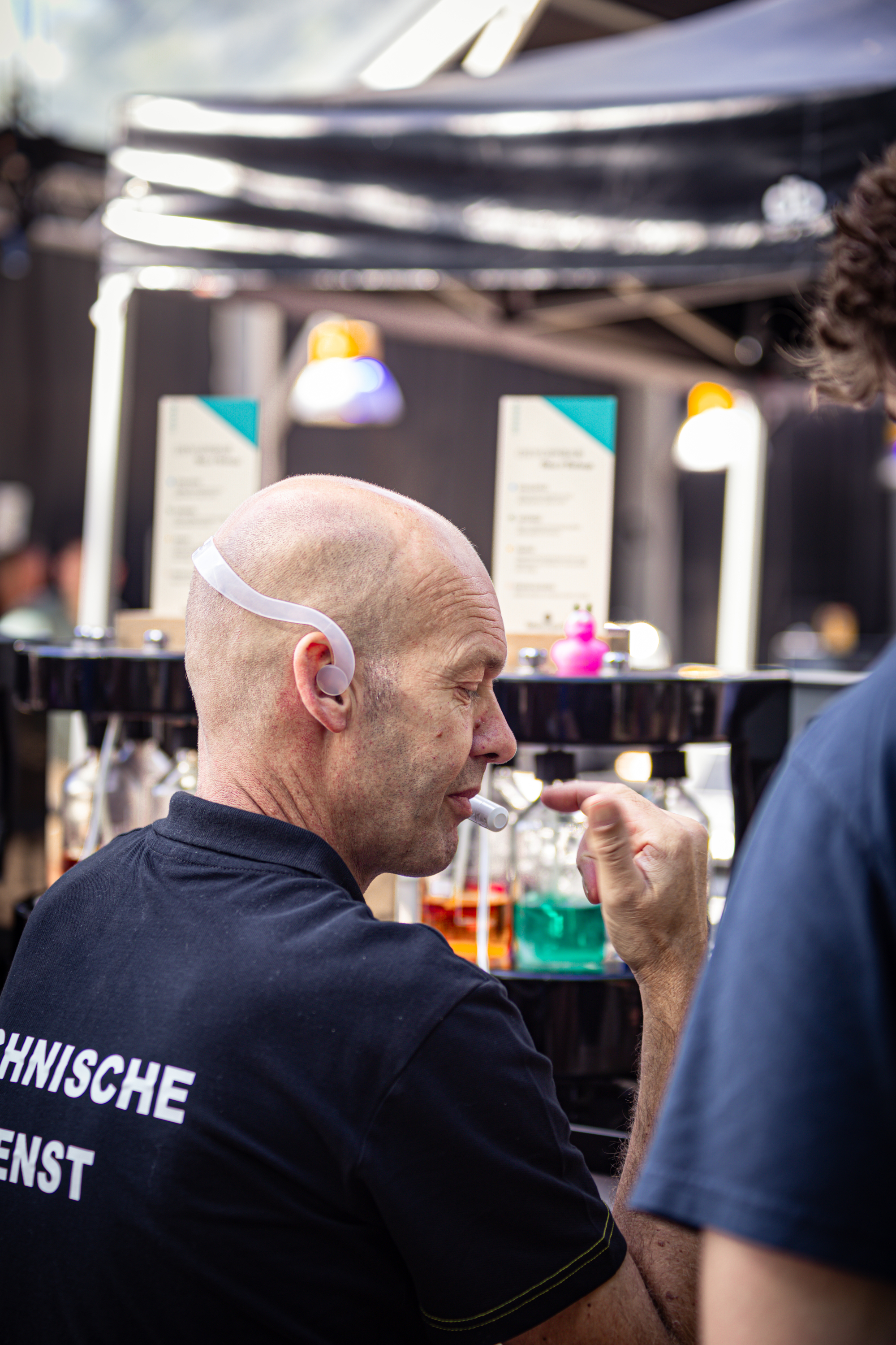 A man standing at a bar with the word Kninsche on his back. He is wearing a hairnet and holding a drink in his right hand.