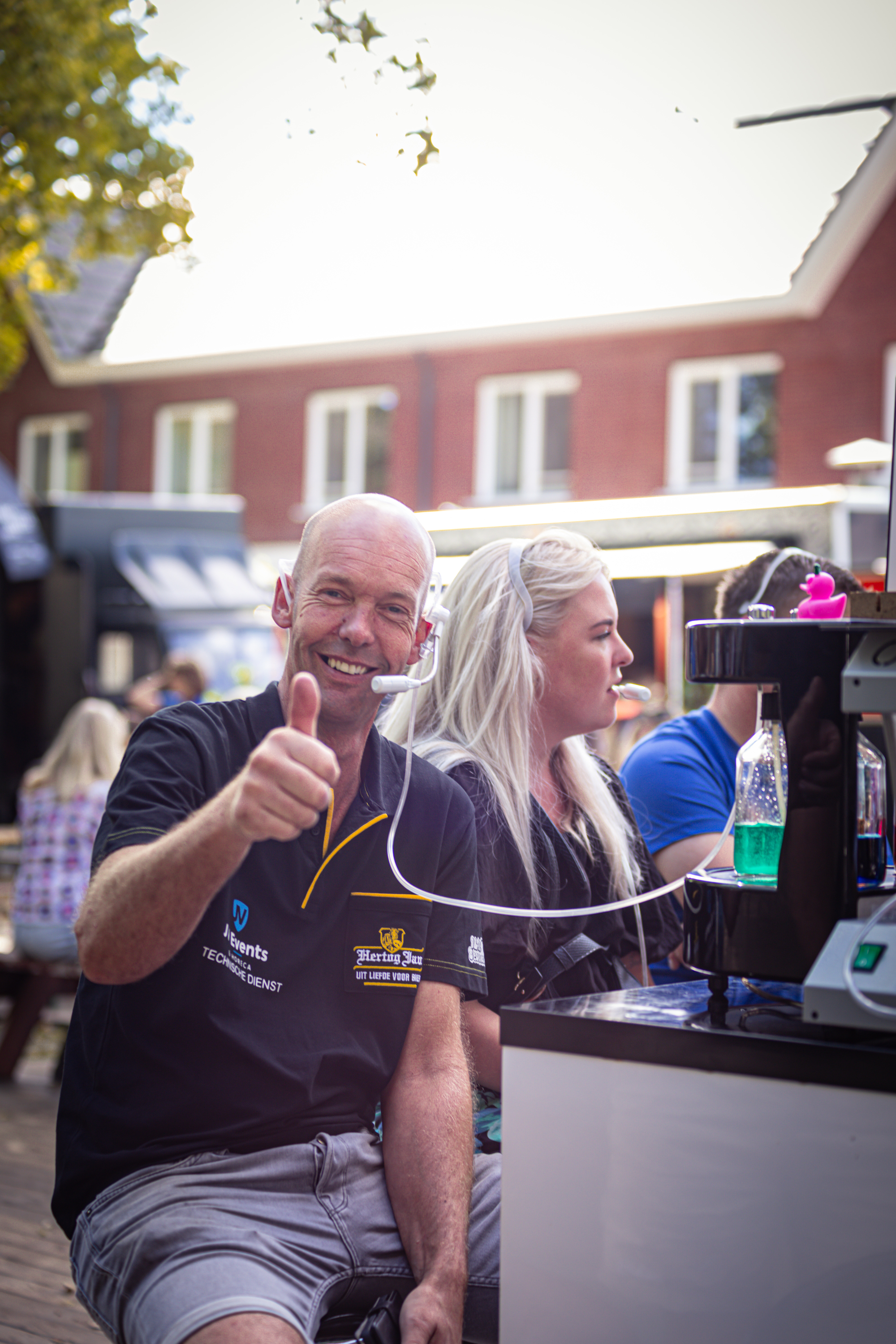 A man is seen with a woman, smiling and giving a thumbs up. They are in front of a coffee shop on a sunny day.