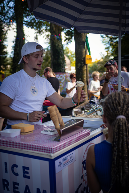 A man wearing a white t-shirt serves a young girl ice cream in an outdoor setting.